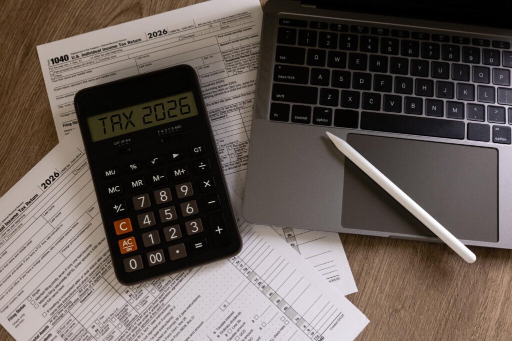 Calculator showing Tax 2026 on top of tax forms beside a laptop